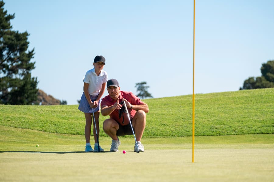 Golf instructor with child student on putting green at golf course lining up putt